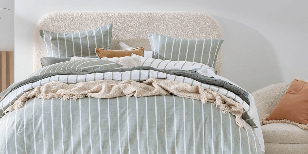 A cozy bed styled with sage green and white striped bedding, a textured cream bouclé headboard, and a wavy woven rattan pendant light hanging above.